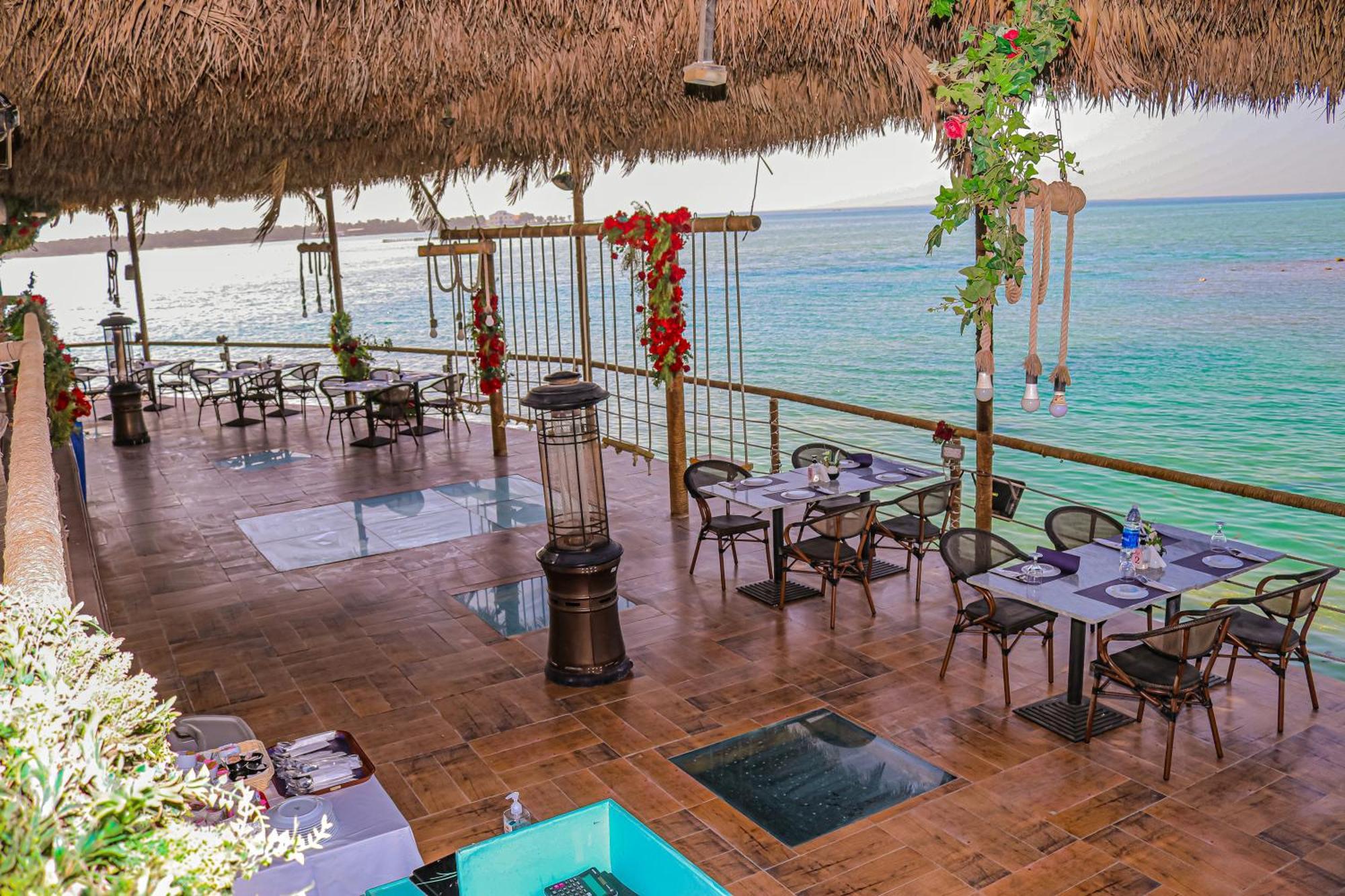 Lagoona Beach Luxury Resort And Spa Манама Екстер'єр фото The photo depicts a coastal restaurant or dining area with an open-air design. It features a wooden floor and tables set for dining, surrounded by chairs. The space is partially covered by a thatched roof, providing shade while allowing for a view of