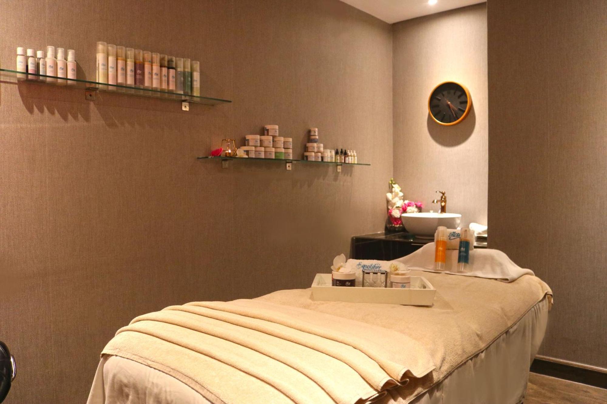 Lagoona Beach Luxury Resort And Spa Манама Екстер'єр фото The photo shows a spa treatment room. There is a massage table draped with a light-colored blanket, creating a relaxing atmosphere. On the wall, there are shelves displaying various skincare products in bottles and jars. A clock is mounted on the wal