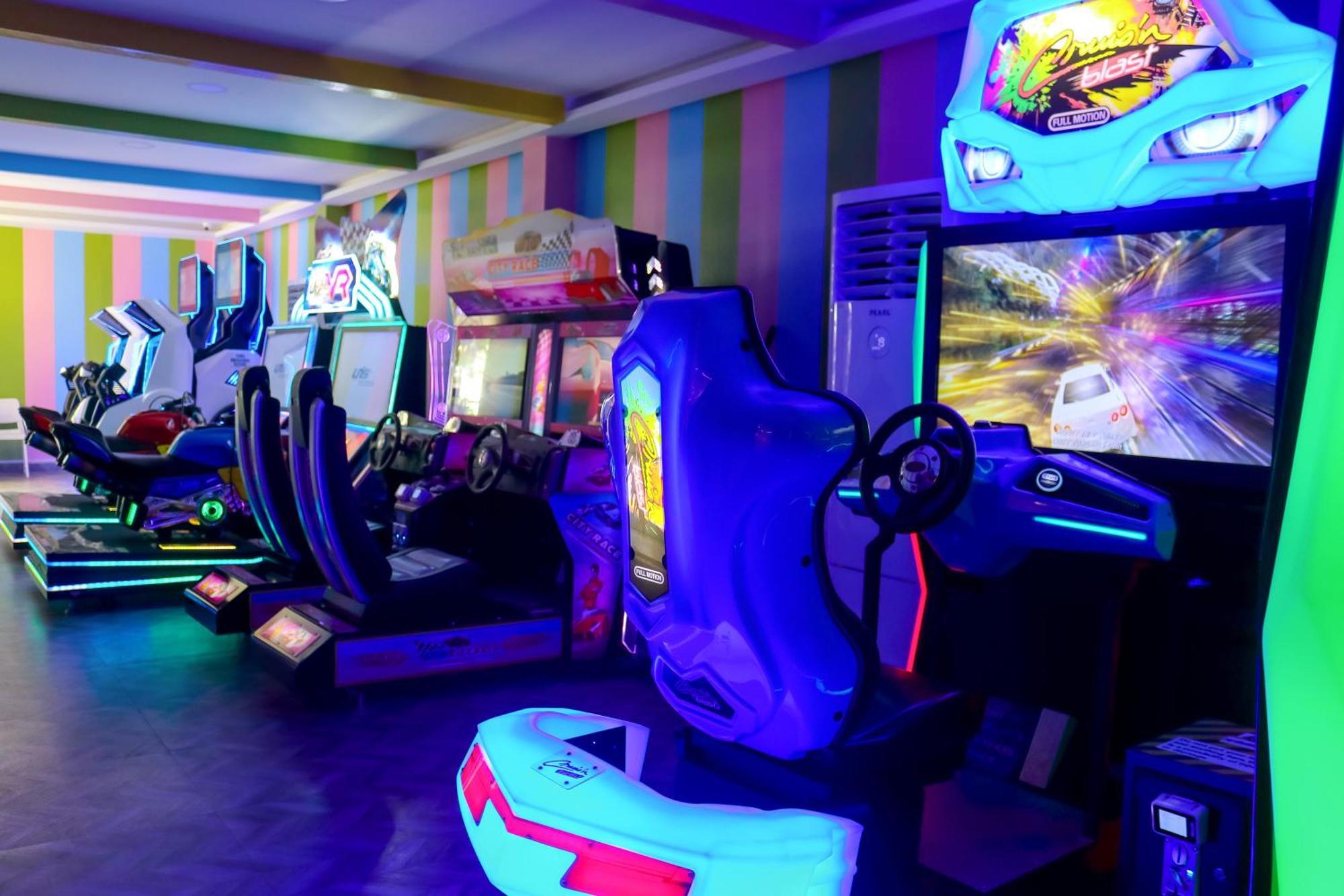 Lagoona Beach Luxury Resort And Spa Манама Екстер'єр фото The photo shows an arcade area filled with various arcade machines. The setting is illuminated with colorful neon lights, creating a vibrant atmosphere. There are several racing game machines with steering wheels and seats, each accompanied by screen
