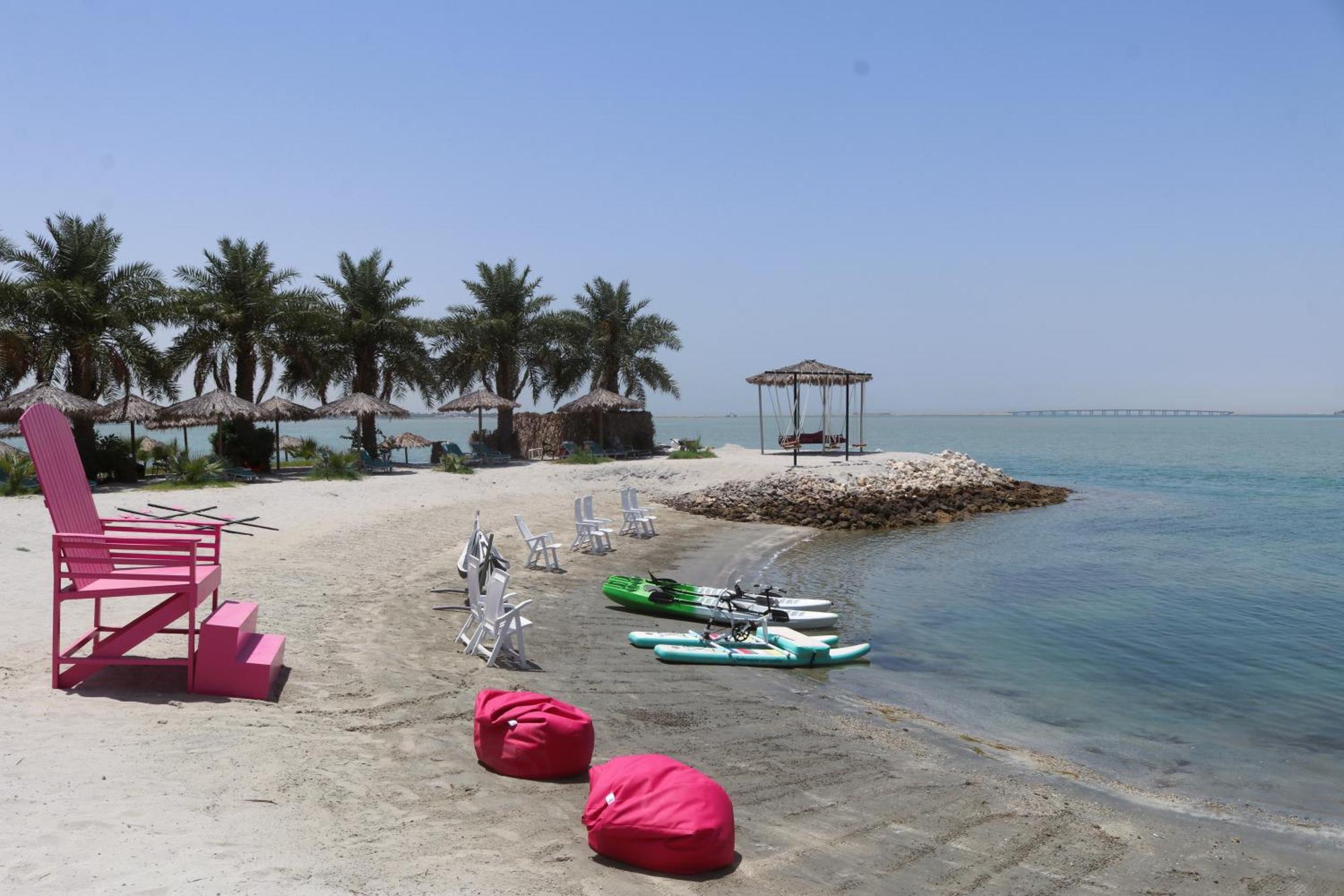 Lagoona Beach Luxury Resort And Spa Манама Екстер'єр фото The photo depicts a serene beach scene with clear blue water and a sunny sky. In the foreground, there are colorful pink bean bags and a large pink chair on the sand. A few white chairs and loungers are positioned near the water's edge. To the side, 
