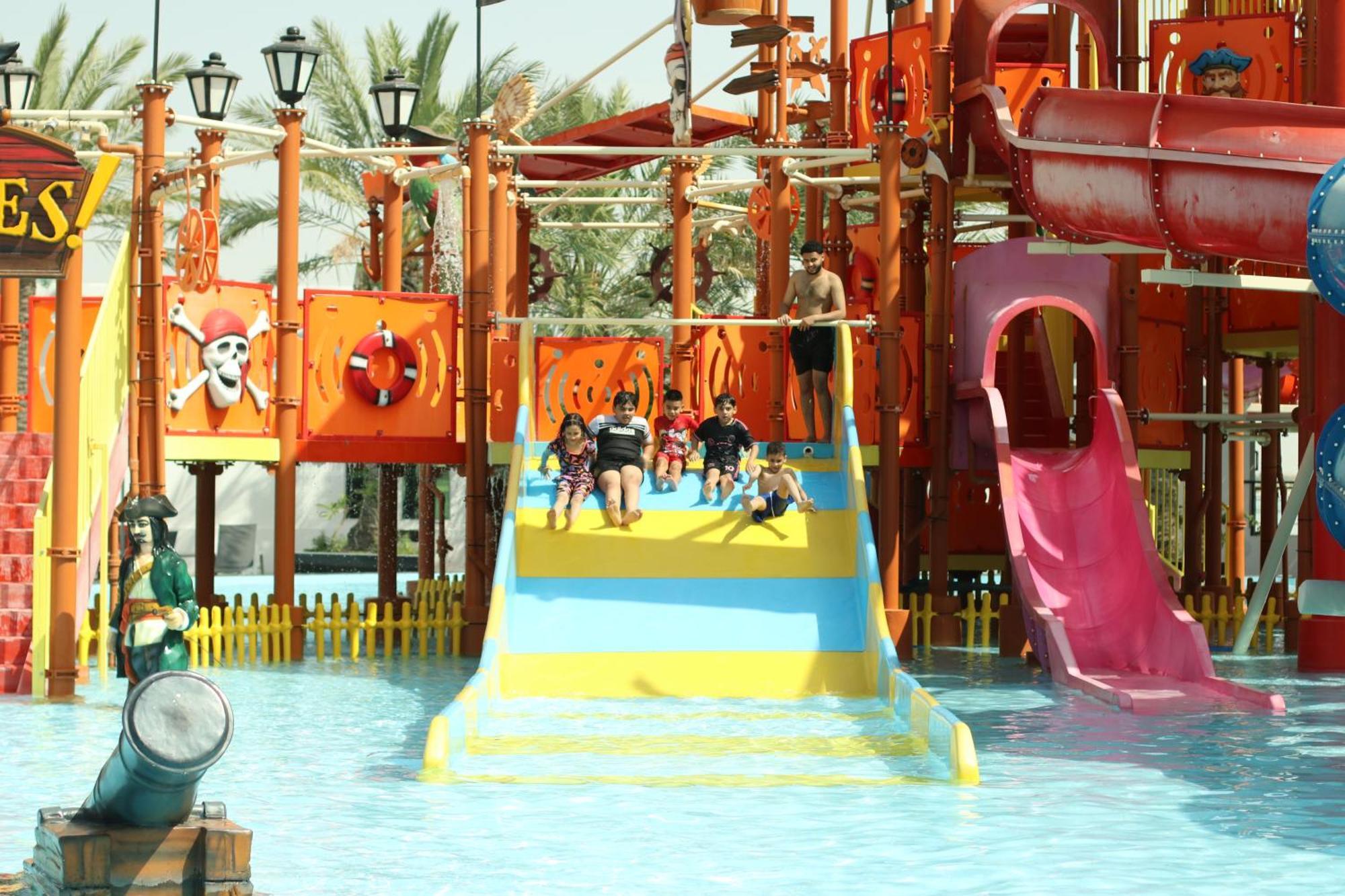 Lagoona Beach Luxury Resort And Spa Манама Екстер'єр фото The photo shows a colorful water park scene featuring a play area with multiple slides. There are children enjoying themselves, sliding down bright yellow and pink slides into a pool of water. The structure has pirate-themed decorations, including a 