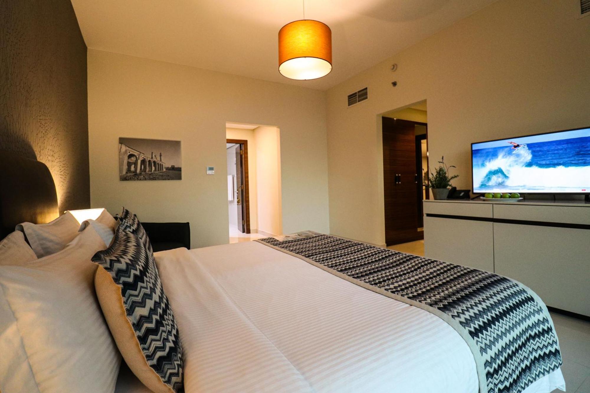 Lagoona Beach Luxury Resort And Spa Манама Екстер'єр фото The photo shows a well-decorated hotel room. There is a large bed with white linens and decorative pillows featuring a zigzag pattern. On the wall beside the bed is a piece of artwork, and there is a modern television on a white furniture unit opposi