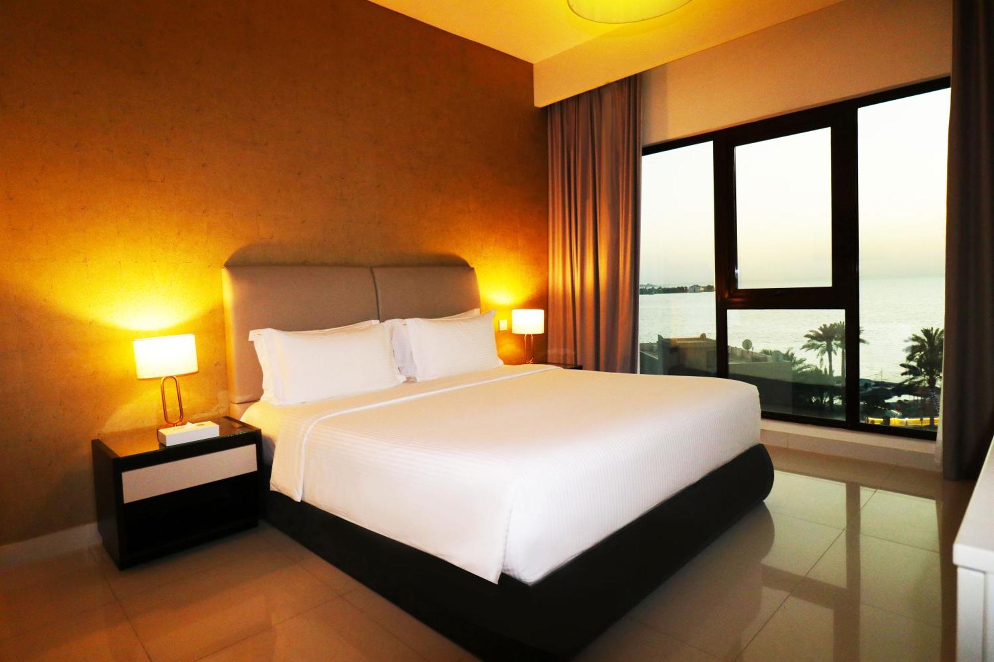 Lagoona Beach Luxury Resort And Spa Манама Екстер'єр фото The photo shows a modern hotel bedroom with a large, comfortable bed dressed in white linens. The room features warm lighting, including bedside lamps, creating a cozy atmosphere. There are large windows with a view of the outdoors, possibly overlook